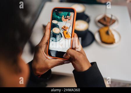 Alto angolo di raccolto anonimo African American femmina blogger registrazione video di caffè tardivo e torte sul cellulare a riunione con amico in esterno c Foto Stock