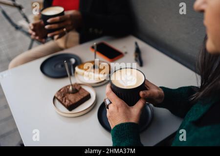 Alto angolo di raccolto anonima femmina con tazza di latte aromatico con arte seduta a tavola con torte durante l'incontro con la ragazza afro-americana Foto Stock