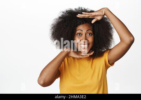 Primo piano di sorridente donna afroamericana che mostra il gesto della cornice della testa, pulito viso luminoso chiaro senza trucco, in piedi su sfondo bianco Foto Stock