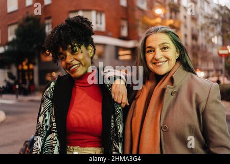 Positiva diverse amiche mature in abiti caldi guardando la macchina fotografica mentre si trova sulla strada della città di sera Foto Stock