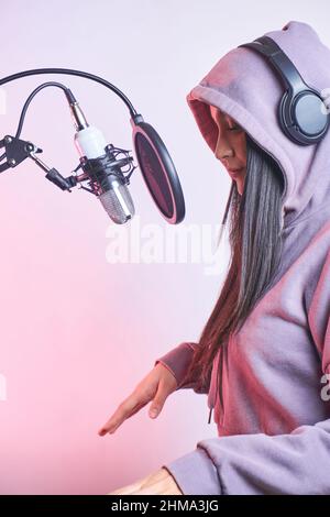 Giovane donna di talento con felpa con cappuccio e cuffie che cantano nel microfono con filtro pop mentre registra la canzone su sfondo viola in moderno stu chiaro Foto Stock