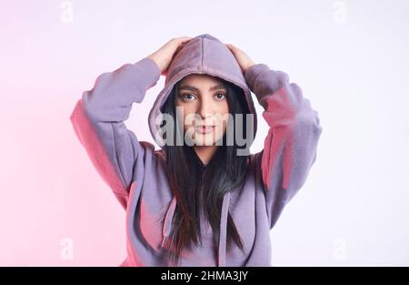 Giovane donna attraente con lunghi capelli neri in felpa con cappuccio casual guardando la fotocamera con sorriso su sfondo chiaro in studio Foto Stock