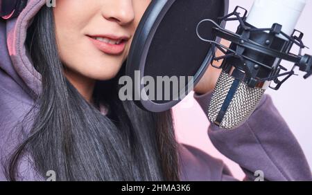 Giovane donna di talento con cappuccio e cuffie che cantano nel microfono con filtro pop mentre registra la canzone sul backgro viola Foto Stock