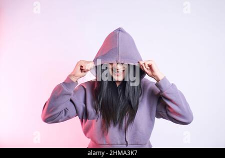 Donna allegra e irriconoscibile con capelli neri che coprono gli occhi con felpa con cappuccio casual mentre si trova su sfondo viola in moderno studio di luce Foto Stock