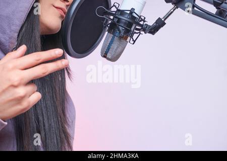 Giovane donna di talento con cappuccio e cuffie che cantano nel microfono con filtro pop mentre registra la canzone sul backgro viola Foto Stock