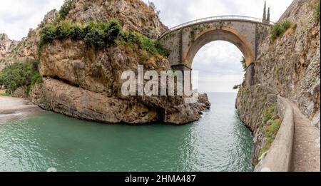 Ampio angolo di vecchio ponte ad arco sul mare ondeggiato situato tra ruvide scogliere rocciose nella natura d'Italia in giorno d'estate Foto Stock