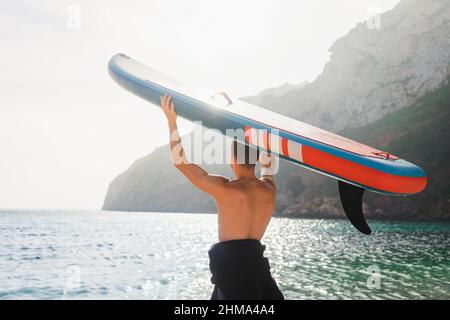 Vista posteriore dell'irriconoscibile surfista maschile con bordo SUP per paddle surf in piedi sulla costa di ciottoli di acqua di mare turchese al mattino Foto Stock