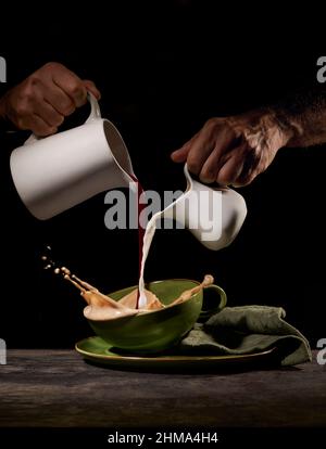 Ritagliare una persona irriconoscibile versando latte e caffè appena fatto in tazza verde sul tavolo in cucina su sfondo nero Foto Stock