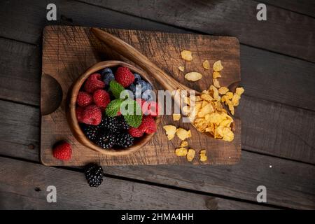 Vista dall'alto del cucchiaio con fiocchi di mais vicino al recipiente con un mucchio di frutti di bosco maturi sul tagliere di legno in cucina Foto Stock