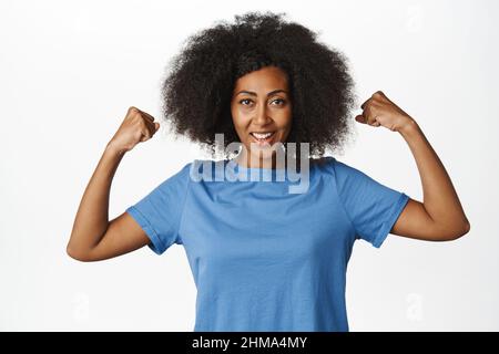 Le donne potere. Entusiasta donna afroamericana che flette bicipiti, mostrando muscoli forti, sentendo forza e guardando fiducioso, in piedi in blu t Foto Stock