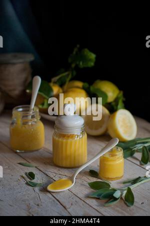 Vasetti piccoli con cagliata di limone fatta in casa gialla posti su tavola di legno con cucchiai e foglie verdi in cucina a casa Foto Stock