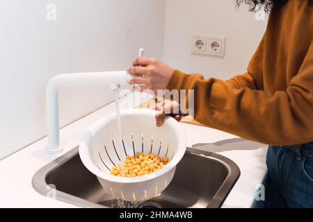 Crop anonima femminile in caldo maglione di lavaggio ceci in colander in lavandino mentre cucinando in cucina a casa Foto Stock