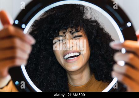Giovane ragazza afroamericana allegra con capelli ricci scuri che ridono mentre illuminata da una lampada ad anello lucida e guardando la macchina fotografica Foto Stock
