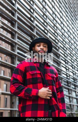 Da sotto serio bearded African American maschio indossando rosso a scacchi camicia e cappello nero in piedi vicino a parete di vetro sulla strada della città e guardando venuto Foto Stock