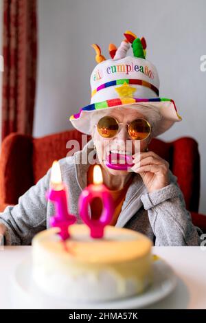 Ragazza anziana allegra in occhiali da sole e divertente cappello mordere candela mentre festeggia il 100 anniversario con torta di compleanno e guardando la macchina fotografica Foto Stock