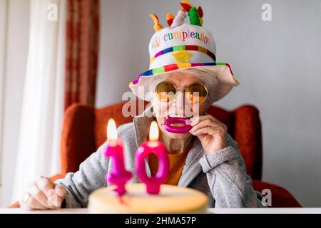 Ragazza anziana allegra in occhiali da sole e divertente cappello mordere candela mentre festeggia il 100 anniversario con torta di compleanno e guardando la macchina fotografica Foto Stock
