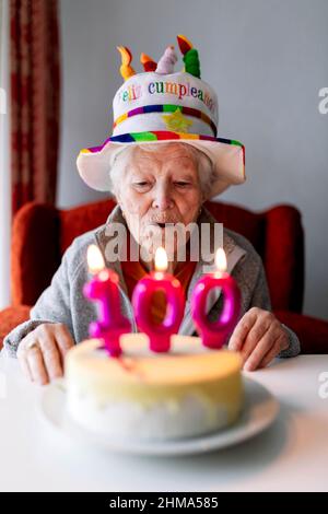 Donna anziana in cappello divertente soffiando candele brucianti con il numero 100 sulla torta di compleanno e celebrare l'anniversario a casa Foto Stock