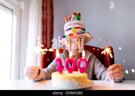 Positiva femmina invecchiata in occhiali da sole e con luci Bengala incandescenti seduti a tavola con torta con 100 candela e celebrare l'anniversario mentre si guarda Foto Stock