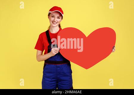 Ritratto di donna operaia ottimista in piedi tenendo il grande cuore rosso nelle mani, guardando la macchina fotografica con espressione felice, indossando tute e cappuccio rosso. Studio interno girato isolato su sfondo giallo. Foto Stock