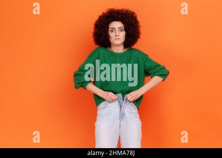 Ritratto di triste giovane donna adulta con acconciatura afroche indossa maglione verde casual stile incapace di buttare jeans a causa di aumento di peso. Studio interno girato isolato su sfondo arancione. Foto Stock