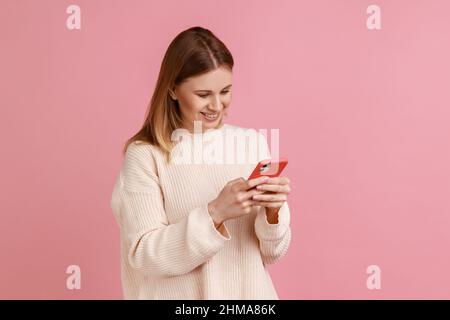 Ritratto di felice soddisfatto bella donna bionda utilizzando smartphone, guardando lo schermo del dispositivo, scrivere post, indossare maglione bianco. Studio interno girato isolato su sfondo rosa. Foto Stock