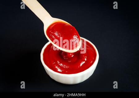 Primo piano di ketchup o salsa di pomodoro rosso in cucchiaio di legno e ciotola bianca su sfondo nero Foto Stock