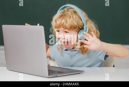 Ragazzo carino che indossa le cuffie, studia con il laptop in aula, ascoltando il corso di lezione audio. Emozioni stupite per i bambini. Foto Stock