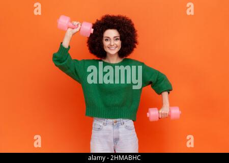 Donna sportiva felice positiva con acconciatura Afro indossare maglione verde casual facendo esercizi per bicipite, utilizzando manubri, assistenza sanitaria. Studio interno girato isolato su sfondo arancione. Foto Stock