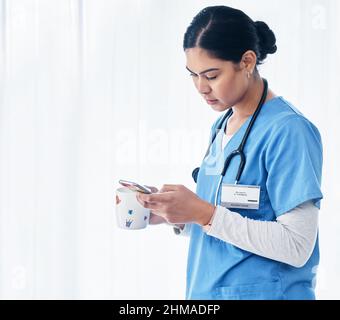 Chatta ora in pausa. Shot di un'infermiera femminile che tiene una tazza di caffè mentre usa il suo cellulare. Foto Stock