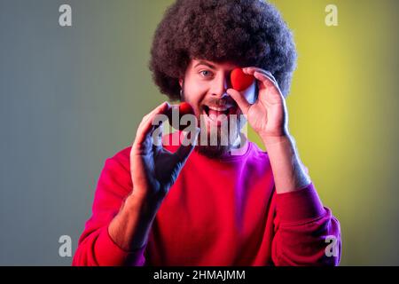 Hipster uomo con acconciatura Afro divertirsi con i cuori giocattolo rossi, mostrando la sua passione e devozione, romanticismo, indossare la felpa rossa. Studio interno girato isolato su colorato sfondo di luce al neon. Foto Stock