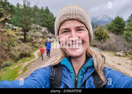 Una mamma felice che prende un ritratto selfie con smartphone in natura in inverno. I suoi figli giocano dietro di lei Foto Stock