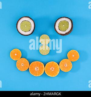 Faccia sorridente creativa fatta di agrumi e cocco su sfondo ciano o blu. Disposizione piatta minima Foto Stock
