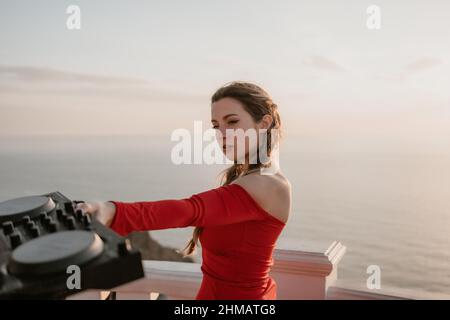 Le mani femminili del DJ suonano la musica mescolando e graffiando durante la festa serale con sfondo marino e caldo tramonto morbido. Primo piano di una console DJ controllata da Foto Stock