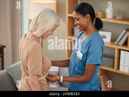 Senza grasso di pancia, Non c’è problema. Shot di un medico che utilizza un nastro di misurazione su uno stomaco donna anziano durante una consultazione a casa. Foto Stock