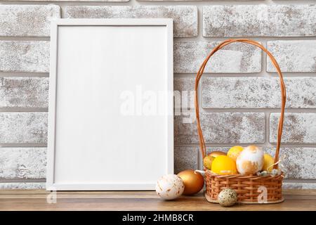 Composizione con cornice fotografica e uova di Pasqua in cestino sul tavolo vicino alla parete di mattone chiaro Foto Stock