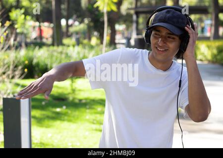 giovane uomo che ascolta musica con le cuffie mentre passeggi nel parco sorridendo e rilassato in mezzo alla natura, indossando un cappuccio e una t bianca Foto Stock