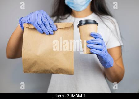 Ragazza consegna cibo lavoratore in una T-shirt dà un ordine in confezioni artigianali con un bicchiere di caffè in una maschera e guanti Foto Stock