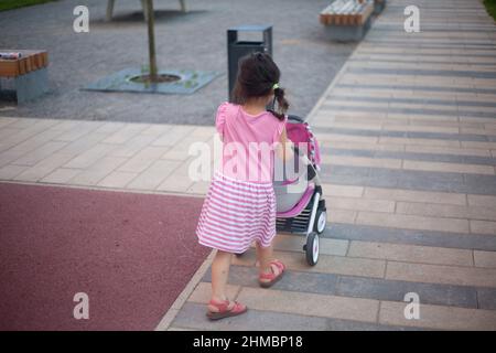 Bambino con passeggino. Ragazza con passeggino per camminare con la bambola. Bambini nel parco giochi. Il preschooler si diverte a giocare all'esterno. Mamma piccola. Foto Stock