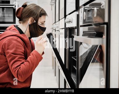 Donna in maschera contro il virus in un negozio sceglie per comprare una stufa da cucina Foto Stock