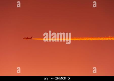 L'aeroplano vola lasciando la traccia di contrail su un alto chiaro nel cielo arancione rosso al tramonto, vista aria-aria Foto Stock