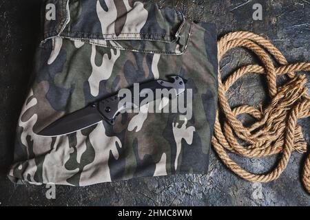 Coltello pieghevole tattico per la sopravvivenza e corda su abbigliamento militare camouflage Foto Stock