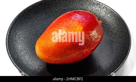 Fresco mango rosso-arancio succoso su un piatto nero isolato su uno sfondo bianco, primo piano Foto Stock