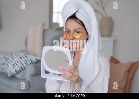 Sotto concetto di trattamento degli occhi e cura del corpo. Bella donna caucasica applica le macchie di bellezza, rimuove i cerchi scuri e puffiness, osserva stupefacente a se stessa nello specchio, siede in una sedia nel soggiorno Foto Stock