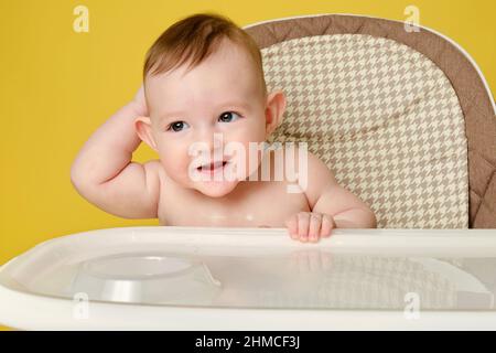 Curioso bambino siede su una sedia alta per l'alimentazione dei bambini, studio sfondo giallo. Bambino ragazzo all'età di sei mesi mangia mentre si siede su un alto chai Foto Stock