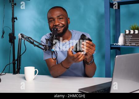 Sorridente revisore di tecnologia afroamericana che presenta le funzioni dell'obiettivo zoom dslr in podcast online dal vivo. Famoso influencer con microfono che esamina le apparecchiature fotografiche di fronte al laptop. Foto Stock