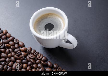 Tazza di caffè bianca con espresso e crema su sfondo scuro con chicchi di caffè sparsi Foto Stock