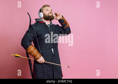 Ritratto di giovane uomo portico in immagine di guerriero medievale, arciere in armatura a catena ascoltando musica in cuffie isolate su sfondo rosa Foto Stock