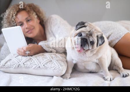 Donna e cane steso sul letto. Gente femminile felice che reding con lettore di ebook e che si rilassa con la sua migliore amica vecchio cane pug. Concetto di anim umano Foto Stock