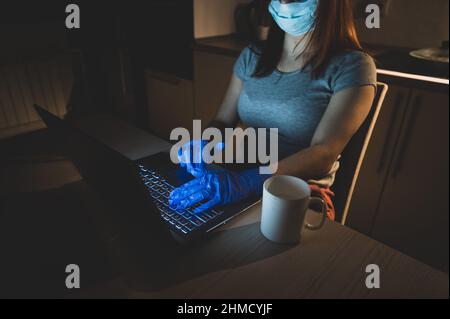 Una ragazza in una maschera e guanti impara durante la quarantena. Una donna che lavora fuori orario da casa su un notebook. Un freelance è seduto in cucina nel Foto Stock
