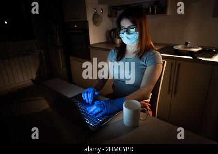 Una ragazza in una maschera e guanti impara durante la quarantena. Una donna che lavora fuori orario da casa su un notebook. Un freelance è seduto in cucina nel Foto Stock